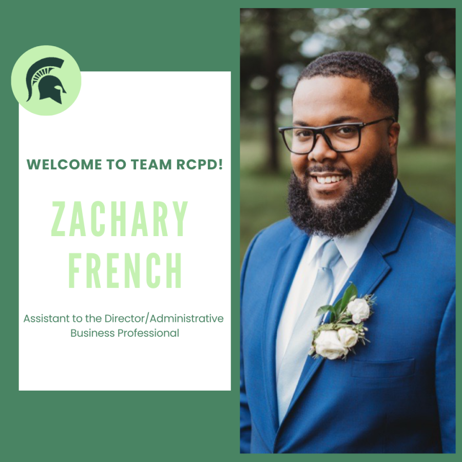 Zach is smiling, has short black hair and facial hair and is wearing a blue suit and tie. He is standing outside.