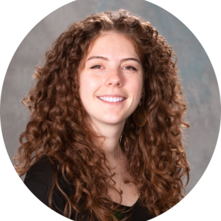 Photo of Kelsey Foote - She is a white woman with long, curly brown hair. She is smiling at the camera and wearing a black shirt.